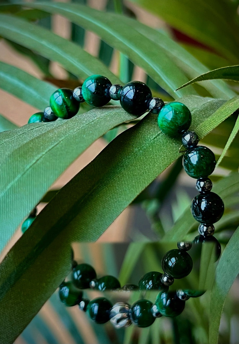 Close-up of a Spirichill bracelet of polished green tiger's-eye and black beads with small hematite spacers, draped over sunlit green palm fronds